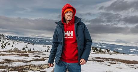 dice «HH» en la sudadera con capucha roja. Persona vestida con una chaqueta de invierno oscura sobre una sudadera con capucha roja y un gorro de punto rojo, de pie en un paisaje montañoso cubierto de nieve