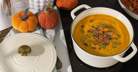 Text reads 'STAUB'. Autumn-themed table setting with white STAUB pot containing orange soup garnished with pumpkin seeds and bacon. Decorative pumpkins, fall leaves, and another pot with vegetables in background.