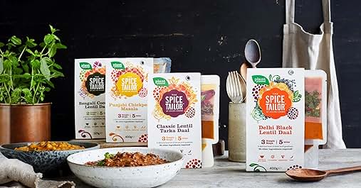 Array of Little Millet food products in colorful packaging displayed with cooking utensils, plant, and ingredients on a dark surface.