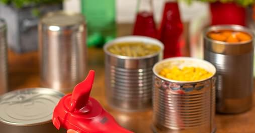Red can opener in foreground. Background shows various canned foods, green glass bottle, red bottles, and leafy green plants in containers on wooden surface.