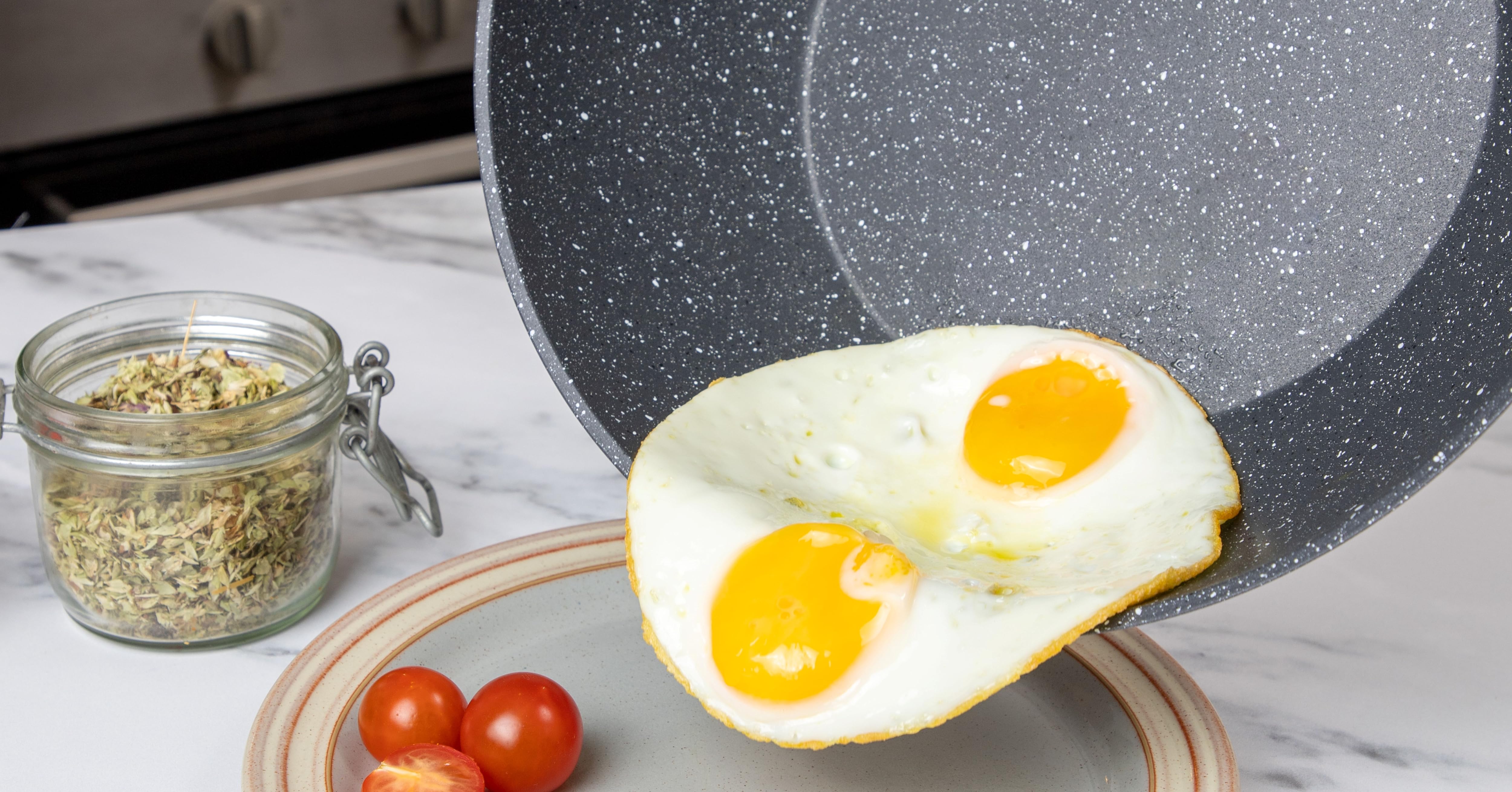 Close-up of a speckled non-stick frying pan cooking sunny-side-up eggs, with wooden utensil and glass jar of seasonings visible.