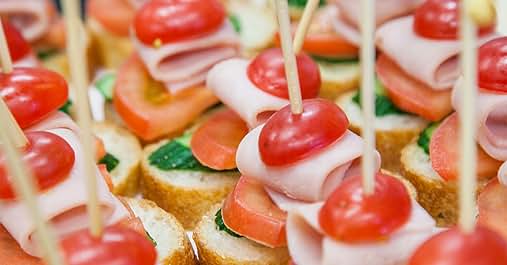 Close-up of appetizers on toothpicks. Small bread slices topped with cheese, ham, cherry tomatoes, and cucumber. Colorful and neatly arranged finger food.