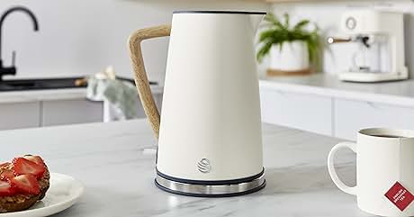 Modern electric kettle in white with beige handle, sitting on kitchen counter with white cabinets and small coffee cup nearby.
