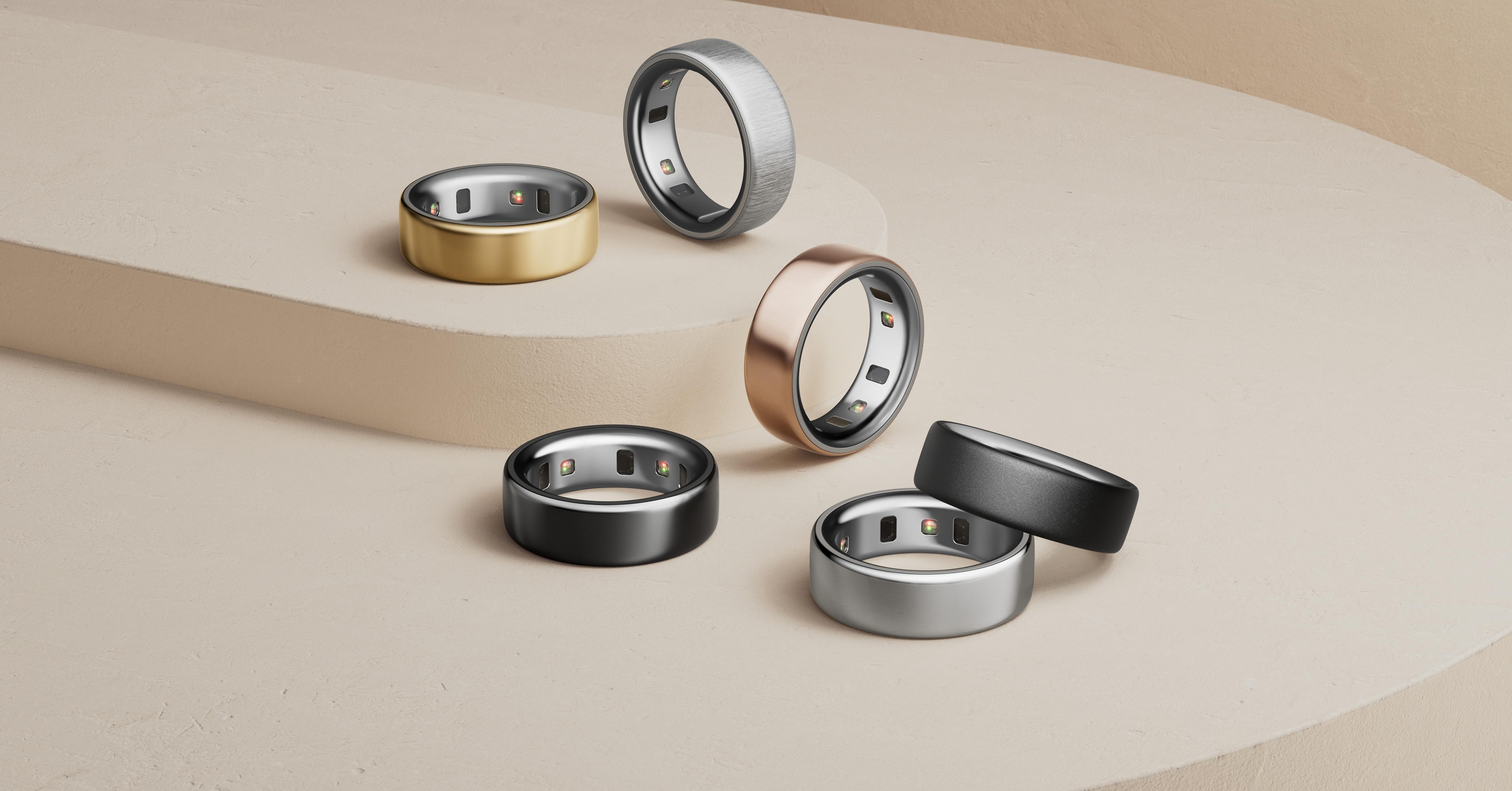 Six stylish metal rings in various finishes including gold, silver, black, and rose gold, arranged on a beige curved surface. Rings feature smooth, wide bands with slightly rounded edges.