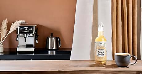 Coffee machine, kettle, and bottle on countertop near curtains. Warm-toned wall background with minimalist setup.