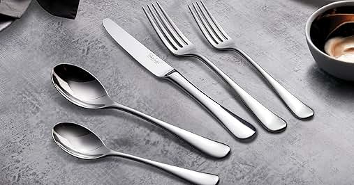 Complete flatware set featuring stainless steel utensils including dinner forks, knife, and spoons arranged on a textured grey surface with ceramic dishes in background.
