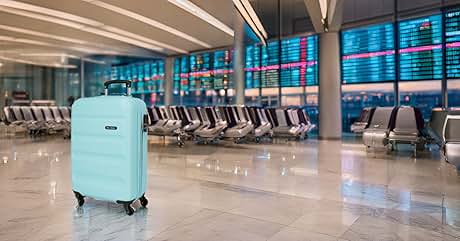 Maleta rígida azul claro con ruedas en una terminal de aeropuerto vacía. Fila de asientos visible en segundo plano