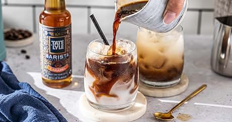 Cold brew coffee being poured over ice in clear glasses, with amber bottle of coffee concentrate visible in background.