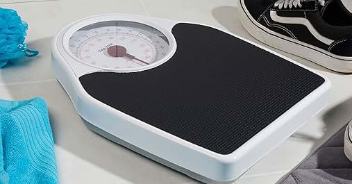 Analog bathroom scale with white base and black non-slip surface. Surrounded by a blue towel and a black sneaker, suggesting fitness context.