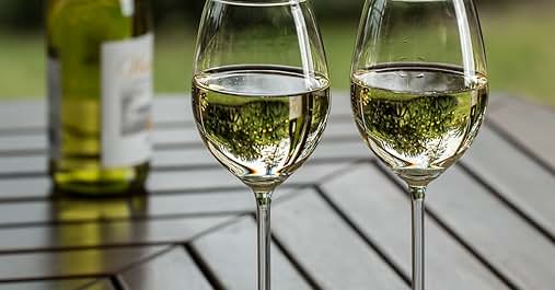 Dos copas de vino llenas que contienen vino blanco sobre una mesa exterior de madera, con una botella de vino parcialmente visible en el fondo.