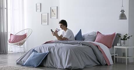 con ropa de cama gris, almohadas rosas y azules, silla blanca y arte mural. Persona sentada en la cama con un teléfono inteligente.