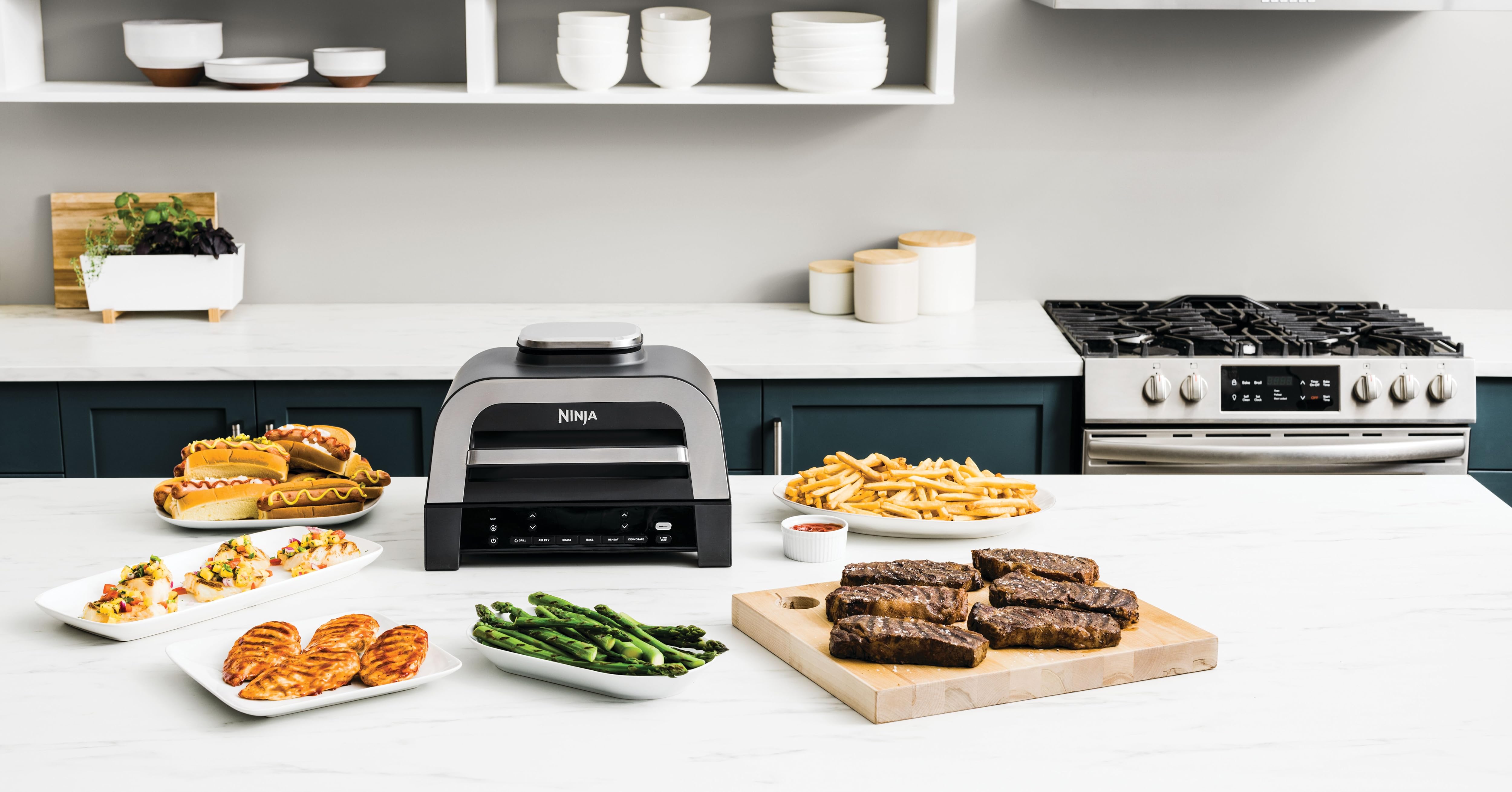 Kitchen countertop with Ooni Karu 16 pizza oven, various prepared dishes including pizzas, asparagus, grilled meat, and bread. White shelves above, stove on right. Showcases versatility of outdoor oven for indoor use.