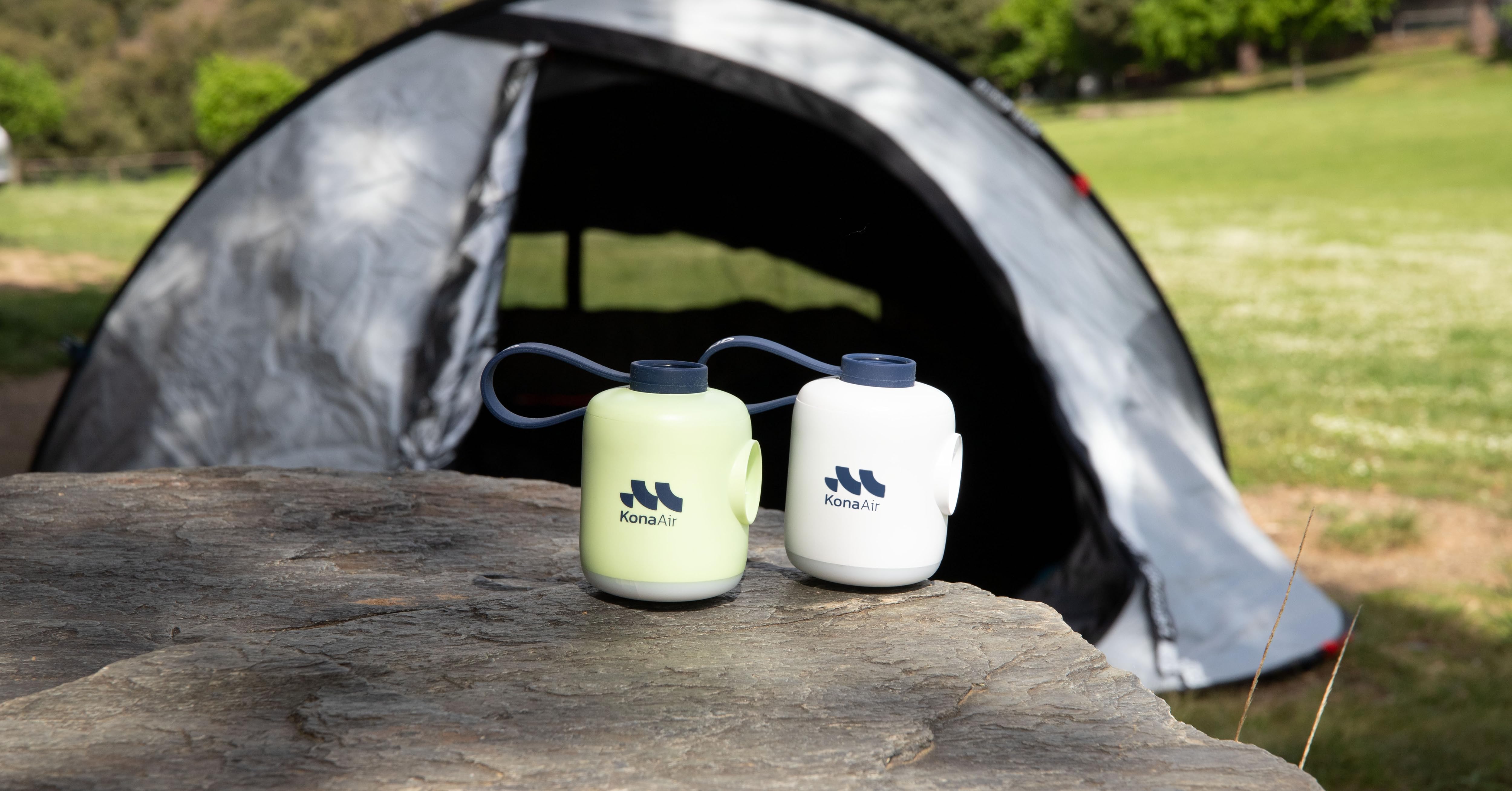 Dos vasos aislantes con logotipos azules se encuentran sobre una roca frente a una carpa gris. Una taza es de color amarillo pálido y la otra blanca. Campamento cubierto de hierba visible en segundo plano