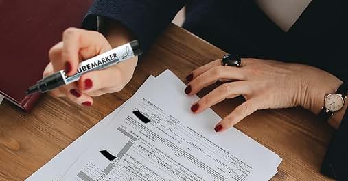Hands on a wooden desk filling out paperwork with a pen, showing part of a document with form fields.