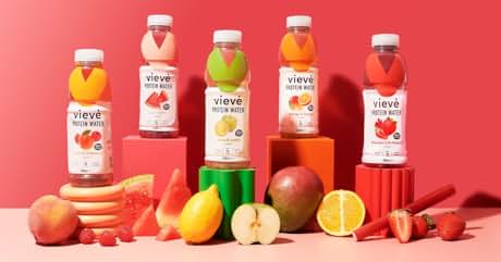 Assortment of fruit juice bottles in white packaging with colorful labels. Fresh fruits like oranges, apples, and strawberries surrounding the bottles on a coral-colored surface.