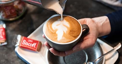 Manos que vierten leche en una taza de café, creando arte con leche. Se ve un paquete de galletas Biscoff en la bandeja