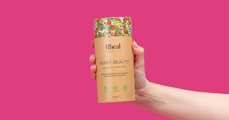 Hand holding a cylindrical cardboard package labeled 'bheal' against a bright pink background. Package appears to contain beauty or wellness product.