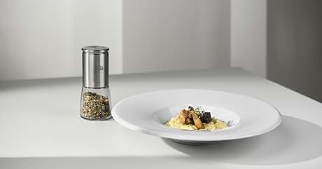 de acero inoxidable con fondo de cristal relleno de hierbas, junto a un plato blanco que contiene una pequeña porción de pasta con verduras.