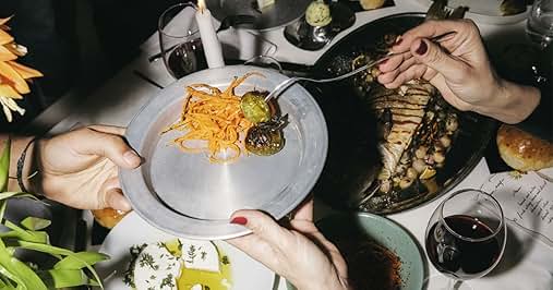 Vista aérea de una mesa de comedor con varios platos de comida. Manos cogiendo platos que incluyen queso rallado, una verdura verde y carne o pescado a la parrilla. Se ve una copa de vino tinto.
