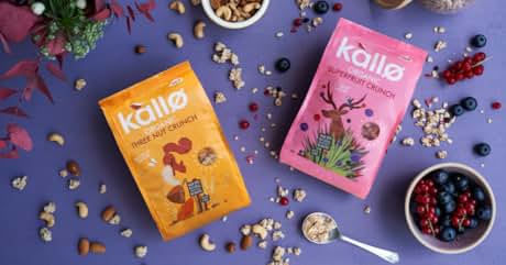 Text reads 'kalló three nut crunch' on orange package and 'kalló super berry crunch' on pink package. Two Kalló cereal boxes surrounded by nuts, berries, and grains on purple background.