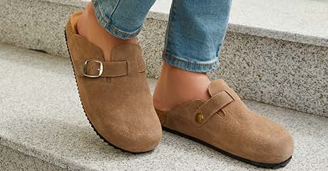 Tan suede clogs with adjustable buckle strap. Person wearing blue jeans and clogs standing on concrete steps. Clogs feature a rounded toe and visible stitching along the edges.