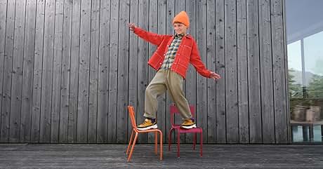 El texto dice «Imagen 1". Persona vestida con una chaqueta roja brillante, un gorro naranja y pantalones caqui que se balancea en dos sillas coloridas sobre una pared de madera como telón
