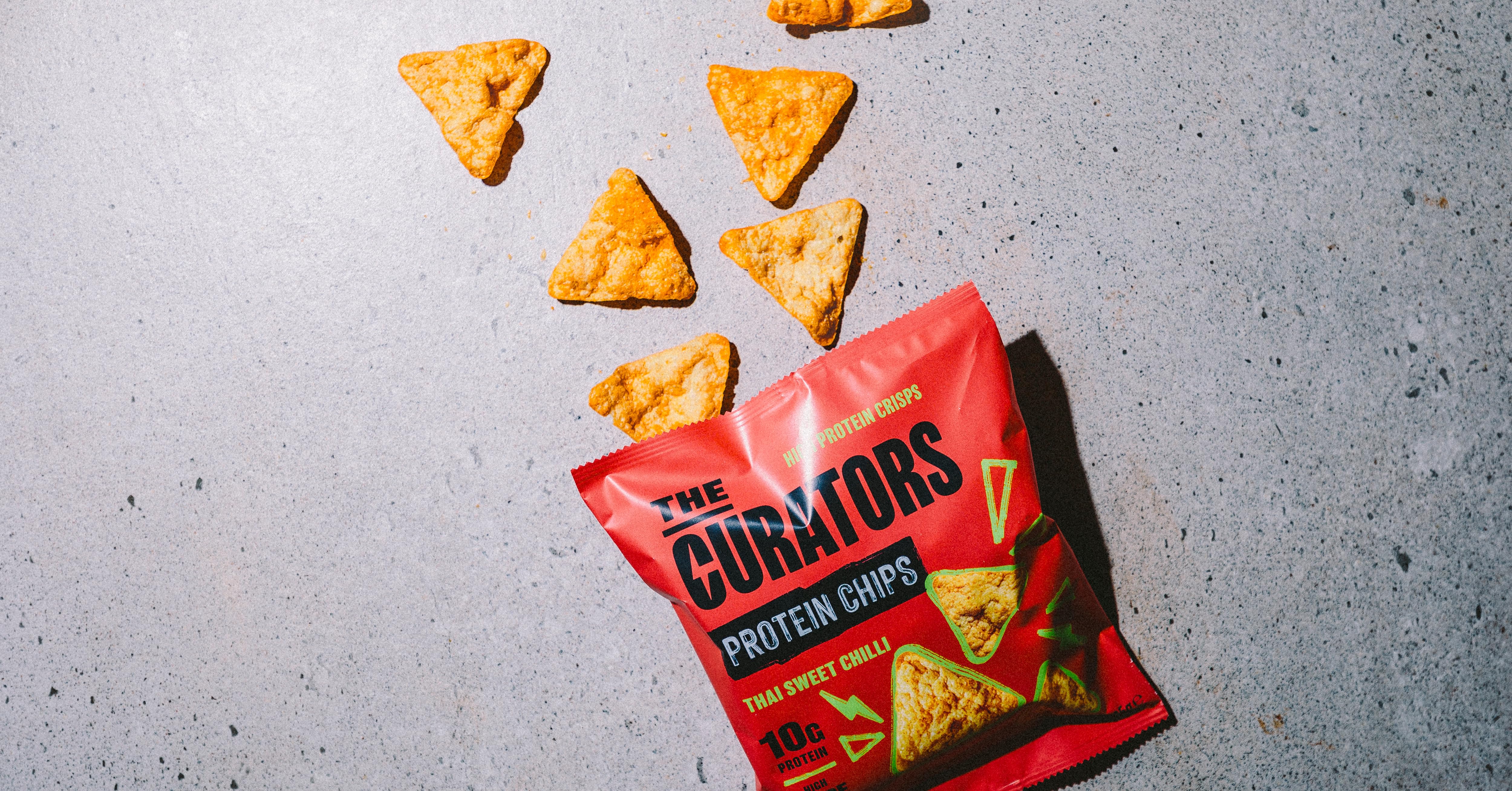 An opened red bag of 'Gurmors Protein Chips' with several triangular-shaped chips scattered on a grey surface. The chips appear crispy and golden in color.