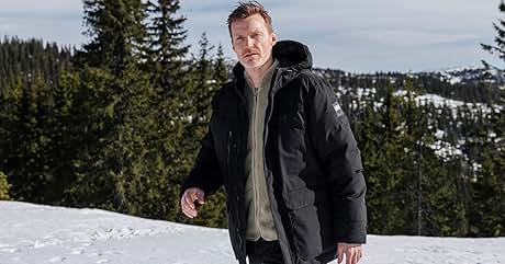 Hombre vestido con una parka negra de invierno con capucha, de pie en un paisaje montañoso cubierto de nieve con árboles de hoja perenne y picos cubiertos de nieve