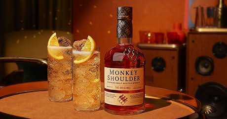El texto dice «MONKEY SHOULDER» en la etiqueta de la botella de whisky. Botella de whisky de color ámbar con dos vasos llenos de hielo y guarnición sobre una mesa de madera en un entorno con poca luz y un equipo de audio de fondo