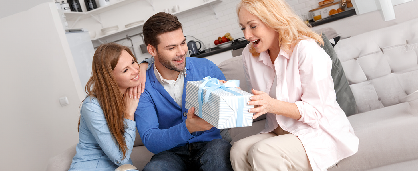 Three people on a couch, two younger individuals presenting a wrapped gift box to an older woman who appears surprised and delighted.