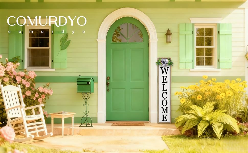 Pastel green house with arched door, &#34;WELCOME&#34; sign, rocking chair, and flower beds.