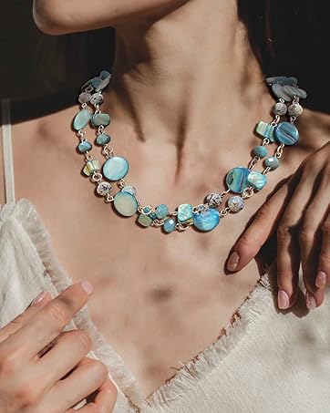 orné de perles circulaires turquoises et blanches de différentes tailles. Le motif à plusieurs brins repose sur la clavicule d'une personne contre un tissu de couleur