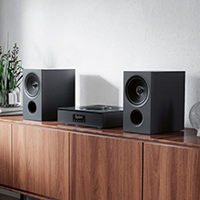Compact stereo system with two black bookshelf speakers and a central unit on wooden cabinet. Speakers feature visible drivers and the main unit has a display.