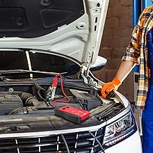 Car with open hood showing engine compartment. Hand in orange glove holds jumper cables near a red portable jump starter device.