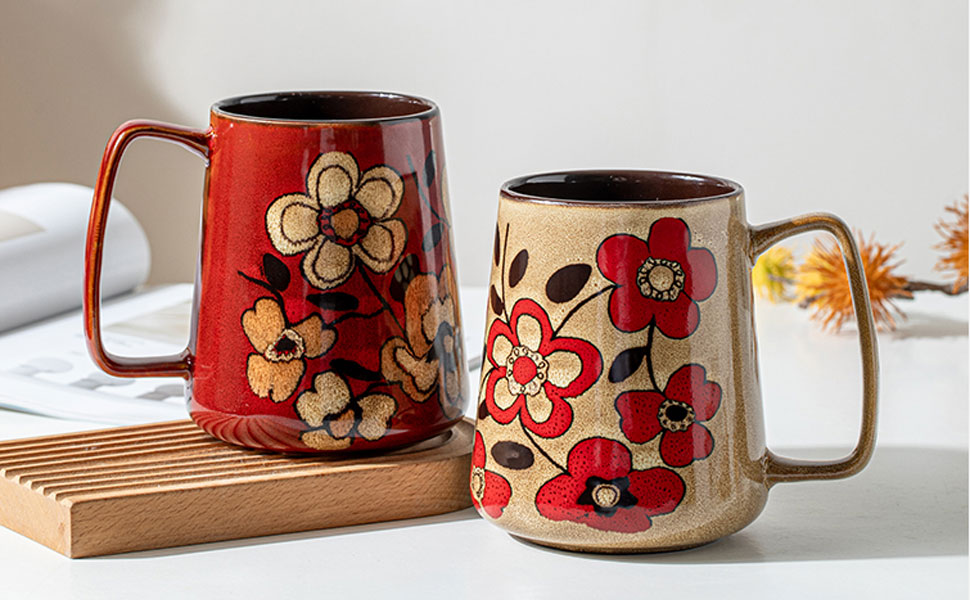 Two ceramic mugs with floral designs. One red, one cream-colored. Both feature large flower patterns and gold-toned handles.