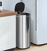Stainless steel cylindrical trash can with a black foot pedal and lid. Placed on a wooden floor next to a white wall.