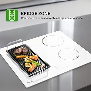 Kitchen cooktop with 'Bridge Zone' feature. White surface with two circular elements and a rectangular grill pan containing meat and vegetables.