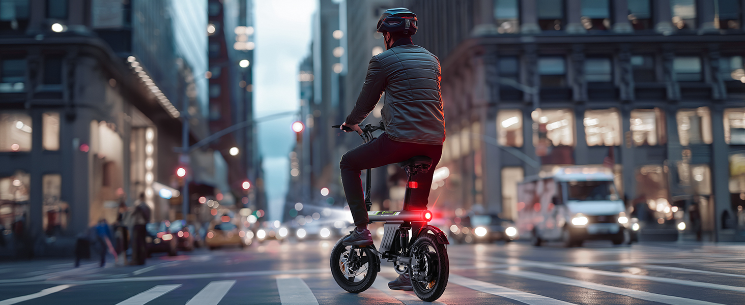 Zusammenklappbares Elektrofahrrad mit rotem Rücklicht, das in der Abenddämmerung in einer städtischen Umgebung mit beleuchteten Gebäuden im Hintergrund gefahren
