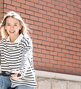 Mujer en top a rayas y jeans, sonriendo mientras sostiene una taza. De pie sobre un fondo de pared de ladrillos, transmite un ambiente informal y relajado
