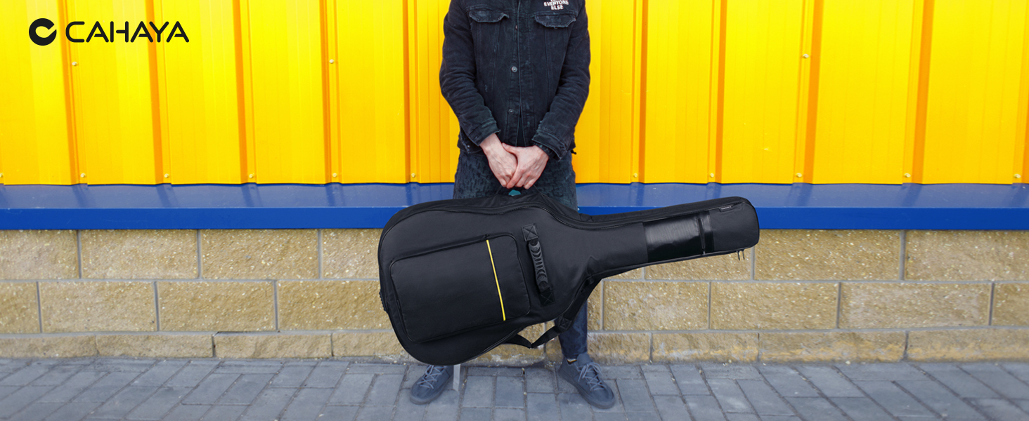Custodia per chitarra imbottita nera con tracolla. Persona in piedi dietro la custodia contro una parete gialla con il logo «CAHAYA» visibile.