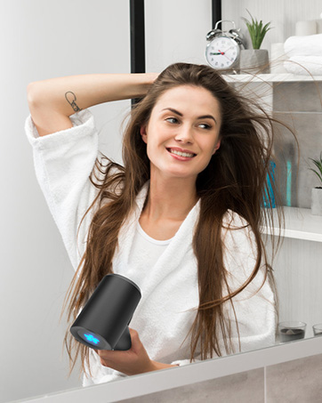 Femme utilisant un sèche-cheveux noir avec un accent bleu dans une salle de bain, entourée d'un décor blanc et d'une petite plante.