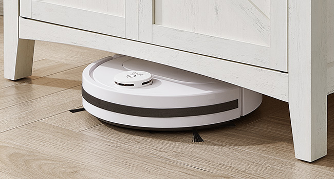 Fehér és fekete kör alakú robotporszívó fa padlón, részben fehér bútorok alatt. Az eszköz alacsony profilú kialakításának köszönhetően szűk helyeken is eligazodhat.
