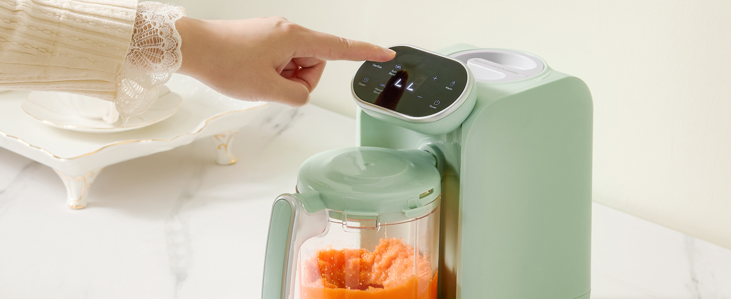 Mint green baby food maker with clear container holding orange puree. A hand is touching the digital display on top of the device.