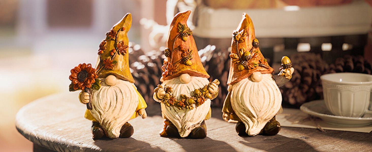 Three decorative autumn gnome figurines with orange pointed hats, white beards, and harvest-themed accessories, displayed on a surface alongside pinecones and a white cup.