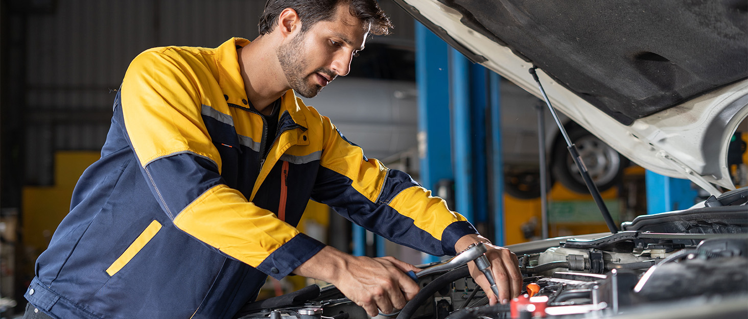 Mechanic in yellow and blue uniform working under a raised car hood in a garage, using tools to perform maintenance or repairs.