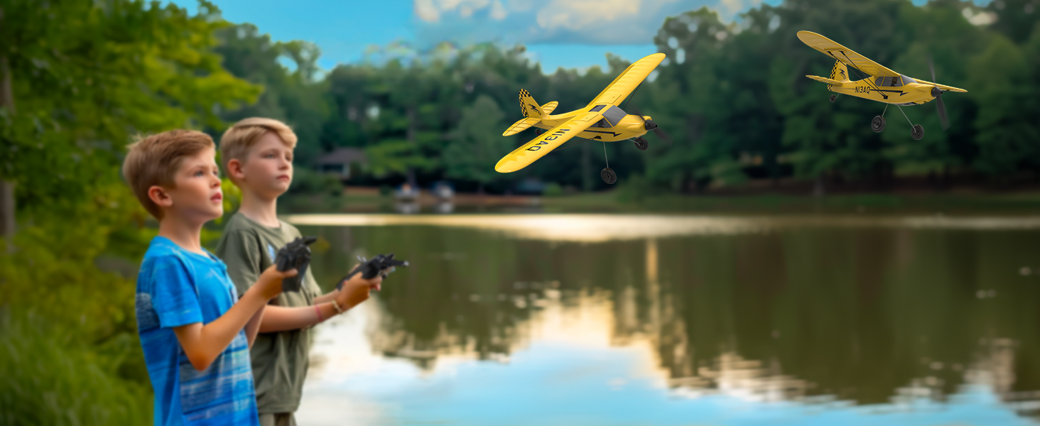 Two yellow remote-controlled toy airplanes flying over a lake while controllers are being operated near the water's edge.