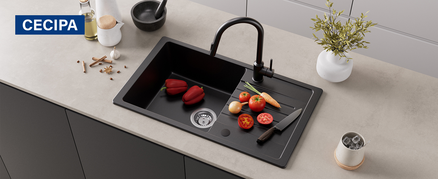 Zwarte spoelbak met kraan geïnstalleerd in lichtgekleurd aanrecht. Rode paprika's en tomaten zichtbaar in de gootsteen, met planten en keukenaccessoires in de buurt.