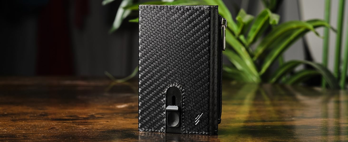 Compact black textured wallet with minimalist design featuring a front pocket and zipper closure, displayed on wooden surface with plants in background.