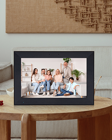 Black digital photo frame on wooden table displaying a group photo of people. Decorative wall art visible in background.