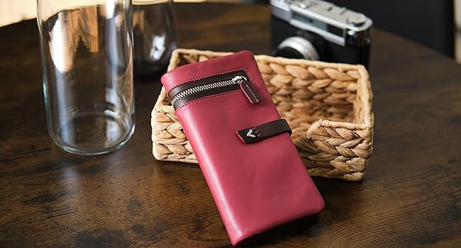 Red leather wallet or case with metallic zipper closure, shown against wooden surface with woven basket and glass container in background.